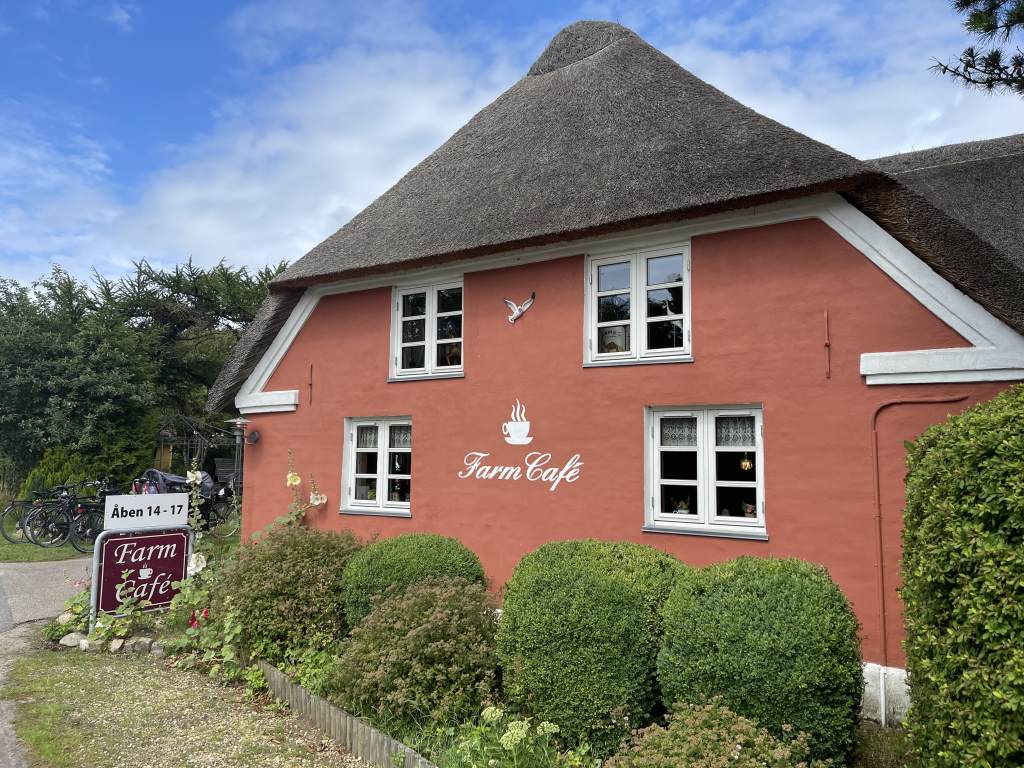 Das Bild zeigt das Farm Cafe, ein charmantes Café mit Reetdach und roter Fassade in Houstrup, umgeben von üppigem Grün.