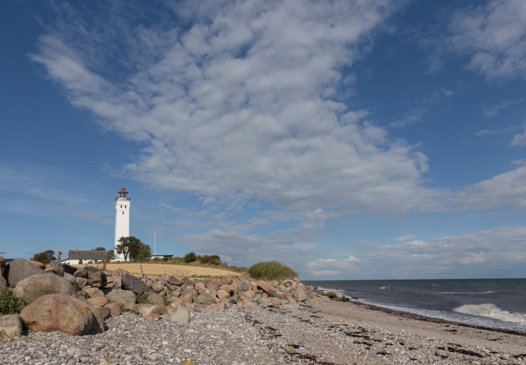 Langeland - Ein weißer Leuchtturm steht majestätisch auf einem steinigen Ufer unter einem dramatischen Wolkenhimmel, wachend über die bewegten Wellen der Ostsee.