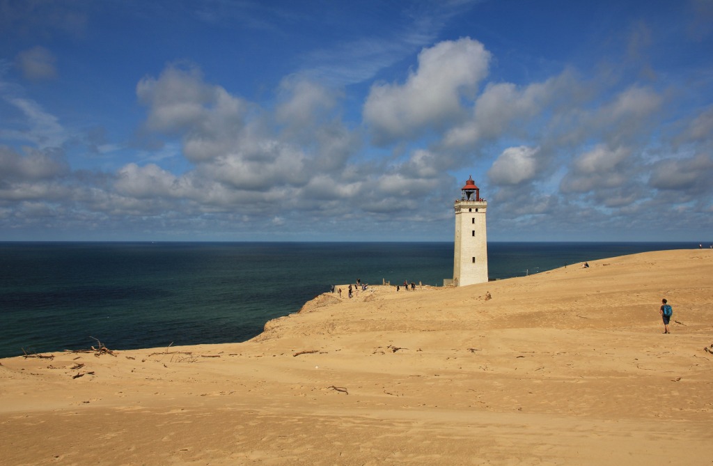 Lökken - Der isolierte Leuchtturm Rubjerg Knude Fyr steht majestätisch an der Kante einer Sanddüne, überblickt das Meer unter einem blauen Himmel mit weißen Wolken.