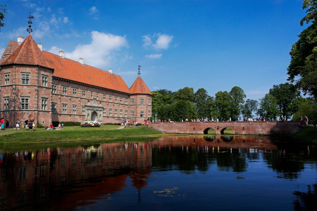 Region Lønstrup - Eine historische Backsteinburg mit Türmchen, umgeben von einem breiten Wassergraben und einer steinernen Brücke, spiegelt sich im ruhigen Wasser.