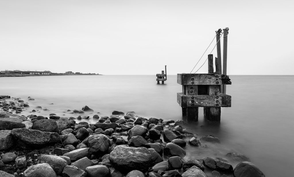 Årgab Hvide Sande - Schwarzweißaufnahme eines alten Holzstegs, der ins ruhige Meer ragt, umgeben von glatten Kieselsteinen am Ufer, unter einem weiten, friedlichen Himmel.