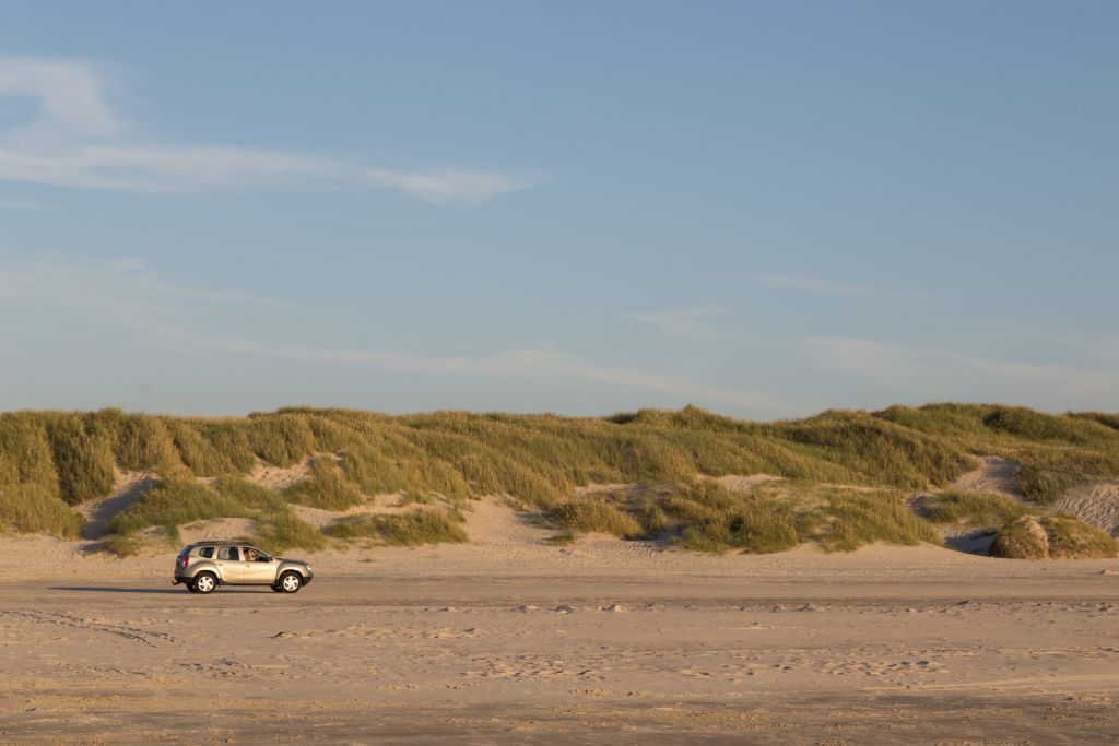 Blokhus Dänemark - Ein einzelnes Auto geparkt am weiten Sandstrand von Blokhus, mit Dünen und Strandhafer im Hintergrund unter einem klaren blauen Himmel in den sanften Farben der Abenddämmerung.