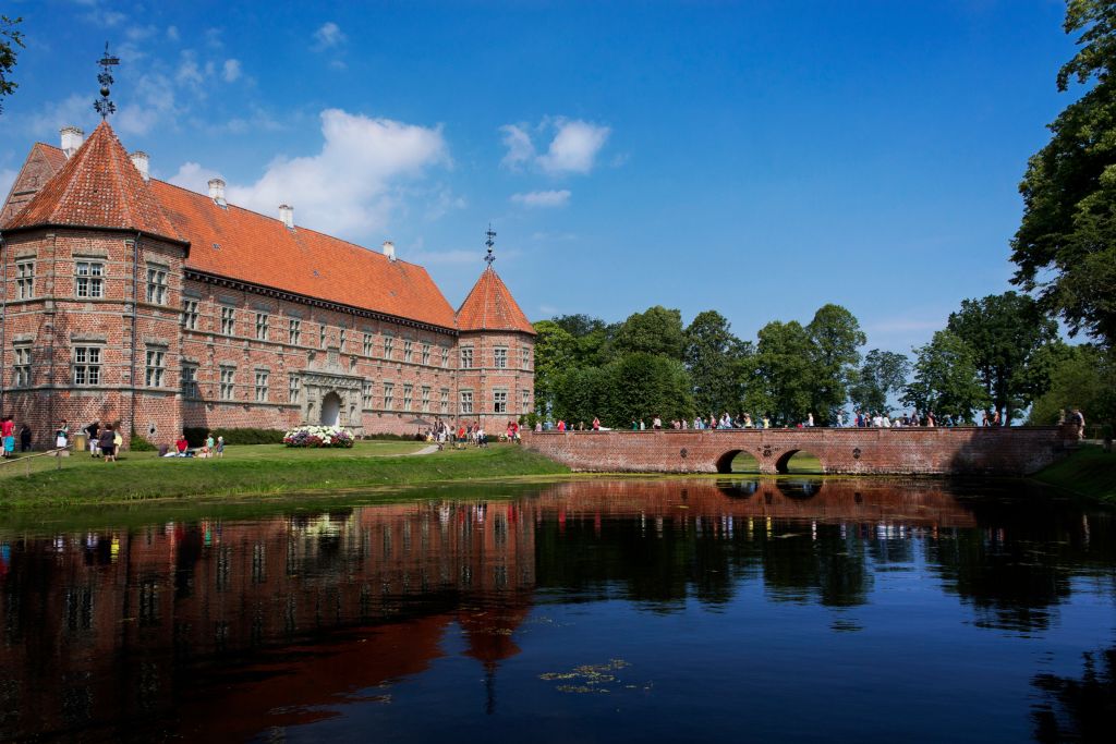Dänemark Blokhus Ferienhaus - Majestätisches rotes Backsteinschloss reflektiert in einem ruhigen Wassergraben, umgeben von grünen Bäumen unter einem blauen Himmel.