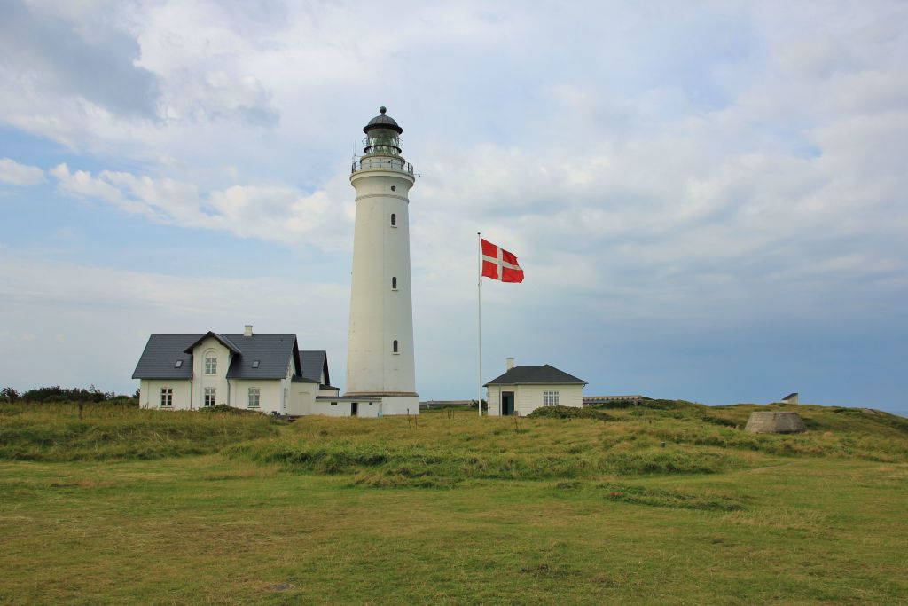 Dänemark Ferienhaus Westjütland - Ein weißer Leuchtturm steht majestätisch auf einer grünen Wiese mit der dänischen Flagge im Wind nahe einem Ferienhaus in Westjütland.