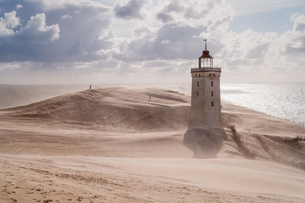 Dänemark Jammerbucht Ferienhäuser - Ein einsamer Leuchtturm, umgeben von sich bewegenden Sanddünen unter einem dramatischen Himmel an der Jammerbucht.