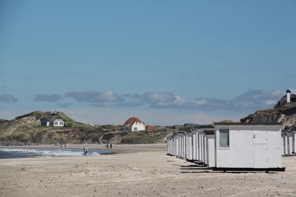 Ferienhäuser Løkken – Weiße Badehütten reihen sich am Sandstrand auf, mit malerischen Häusern auf den Dünen im Hintergrund, ein typisches Bild der entspannten Urlaubsatmosphäre in Løkken, Dänemark.