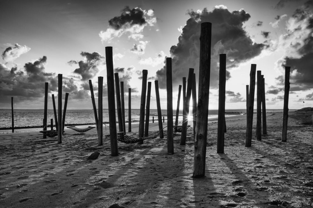Ferienhäuser Westjütland Dänemark - Dramatische schwarz-weiße Darstellung von Holzpfählen am Strand bei Sonnenuntergang, die einst Teil einer Struktur waren, in Westjütland, Dänemark.