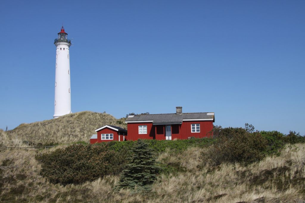 Ferienhaus Årgab Dänemark - Ein charmantes rotes Ferienhaus neben einem eindrucksvollen weißen Leuchtturm, eingebettet in die natürlichen Sanddünen von Årgab unter einem klaren blauen Himmel.