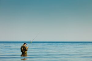 Meeresangeln in Dänemark - Ein Angler in Tarnkleidung steht ruhig im flachen Wasser des Meeres und wirft seine Angelrute aus, umgeben von der Weite des ruhigen blauen Wassers und dem klaren Himmel.