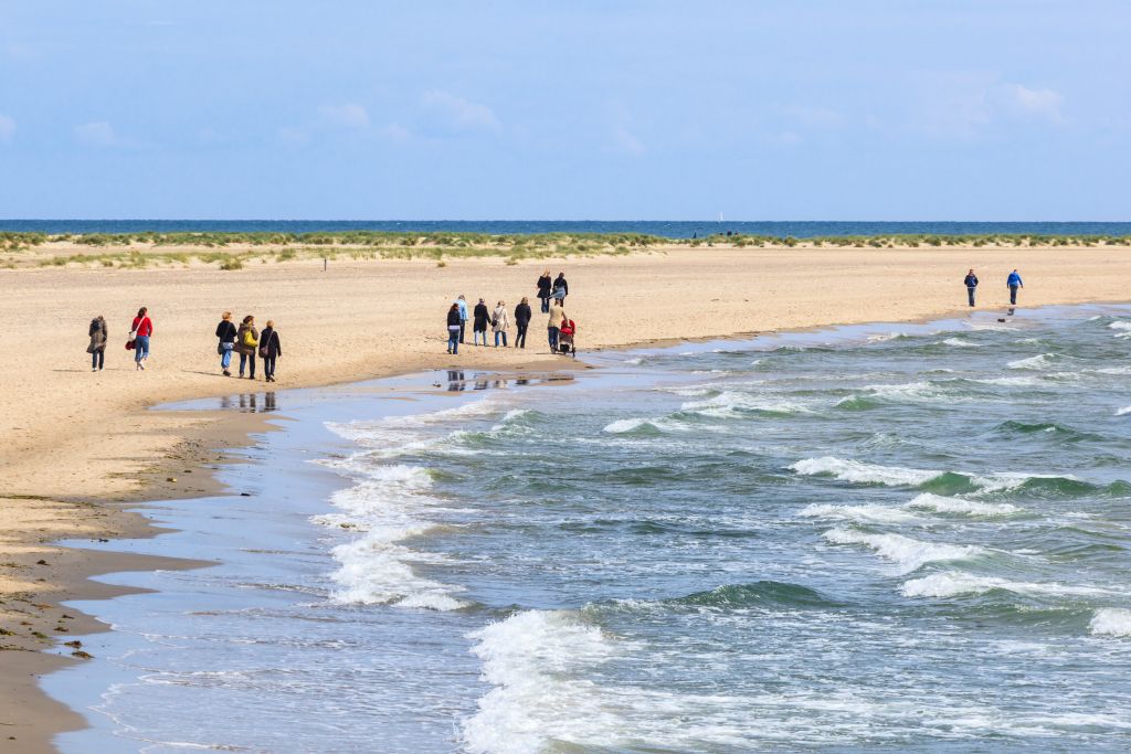 Skagen Dänemark – Besucher schlendern entlang des breiten Sandstrandes von Skagen, umgeben von dynamischen Wellen und einer weiten Dünenlandschaft unter einem hellblauen Himmel.