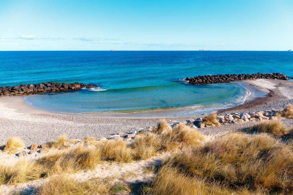 Skagen Denmark – Blick auf eine ruhige Bucht mit klarem türkisfarbenem Wasser, umgeben von steinigen Wellenbrechern und einem sandigen Strand mit Dünengras in Skagen.