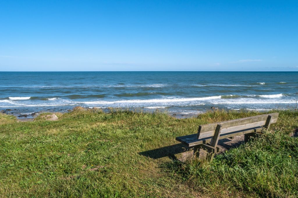 Wetter Hirtshals zeigt einen ruhigen Tag am Meer mit sanften Wellen, blauem Himmel und einer einladenden Bank am Strand, die zur Erholung einlädt.